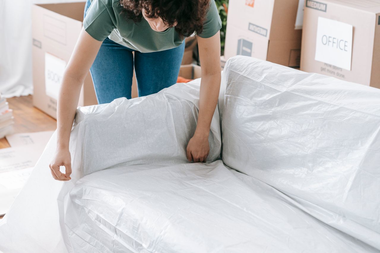 about-us A woman carefully wraps a sofa in protective material while packing boxes in a bright room.